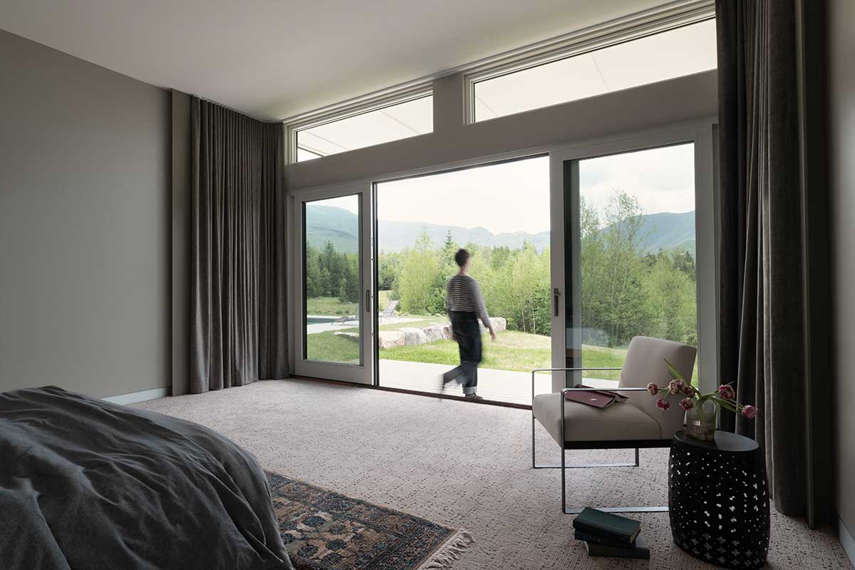 A person in a bedroom walking through a large Marvin Ultimate Sliding French Door to a patio that faces mountains and trees in New Hampshire.